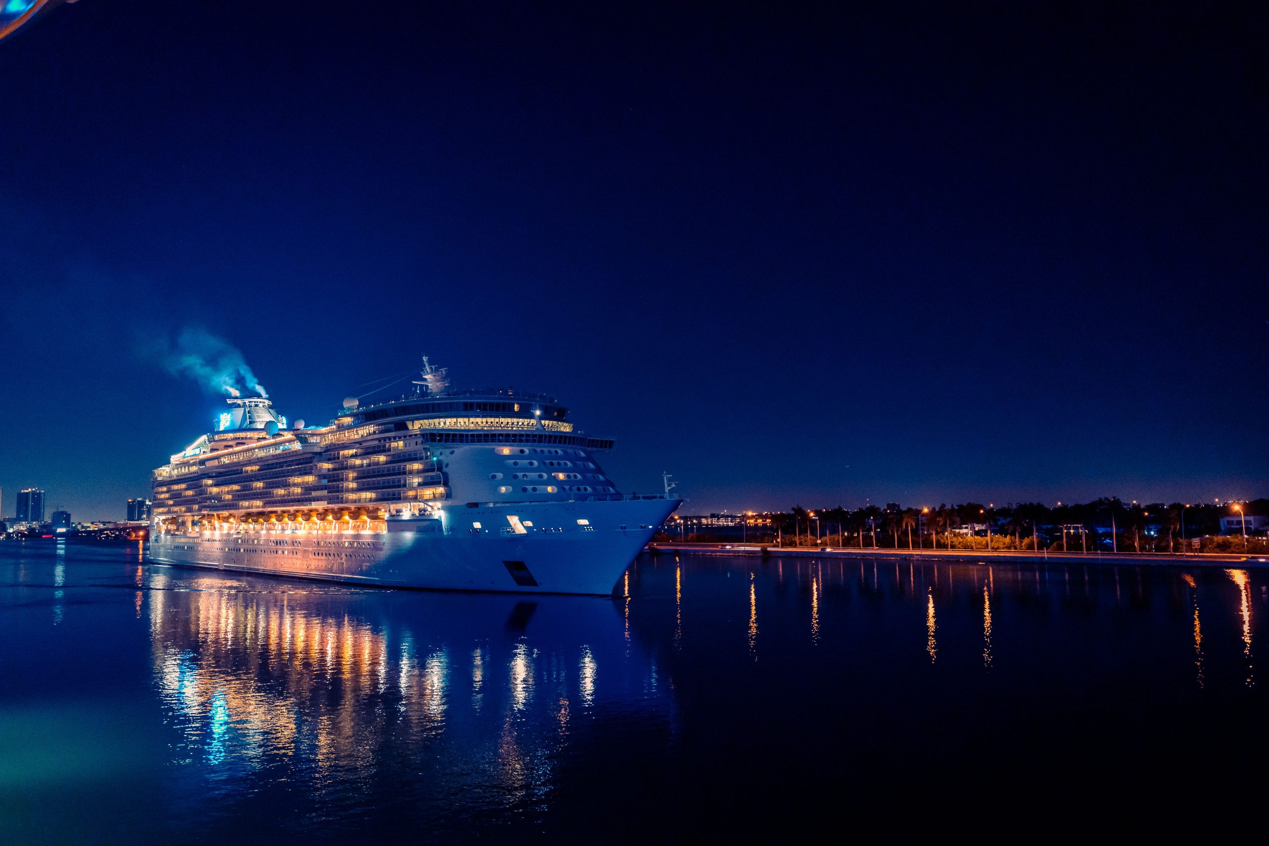 A brightly lit luxury cruise ship sails through Miami's waters at night, reflecting in the calm sea.